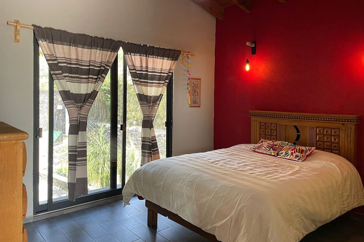 A bedroom with a bed, a wooden headboard, a red wall, and a window with curtains.
