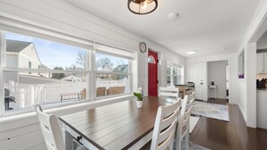 A dining area with a wooden table and white chairs, a red door, and a view of a white picket fence and neighboring houses. - Beach Cottage, private beach and pass (East Lyme)