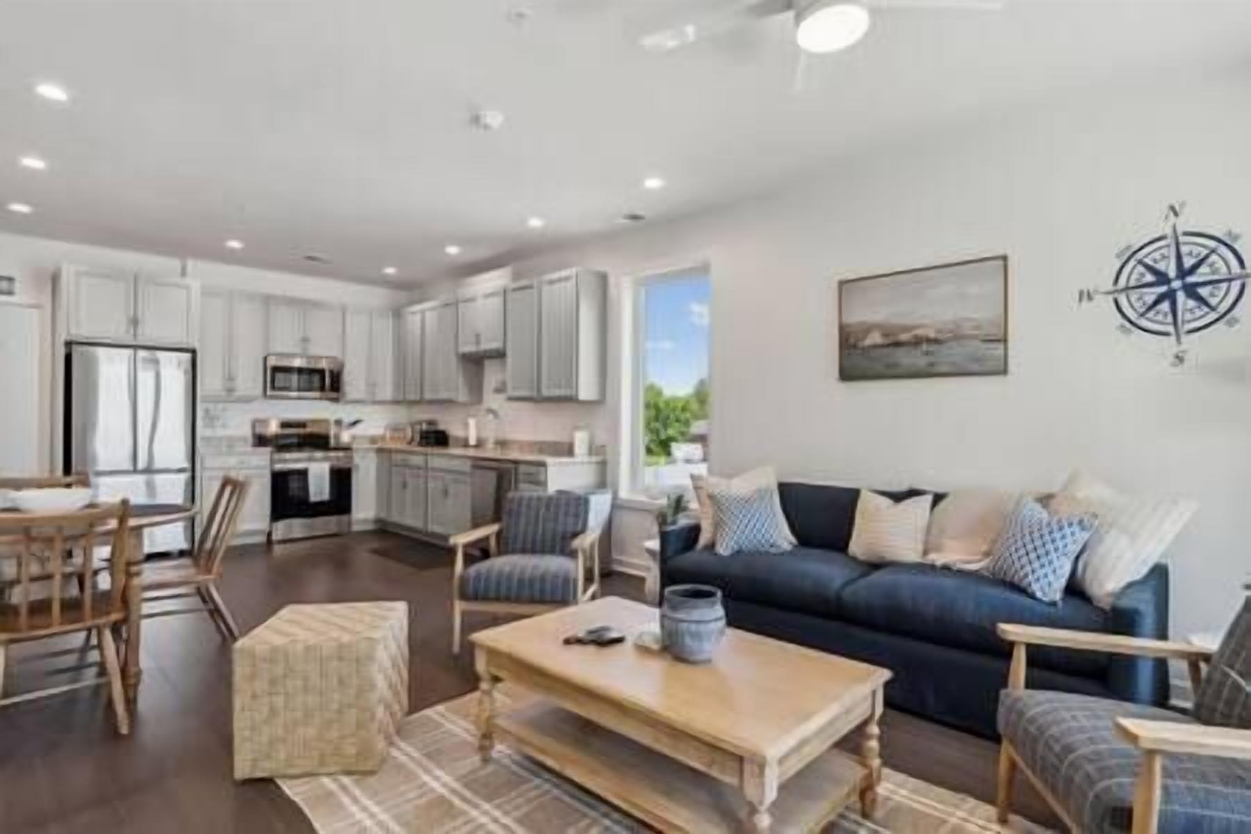 A modern kitchen with white cabinets, a dining area with a wooden table and chairs, and a living room with a dark blue sofa and patterned cushions.