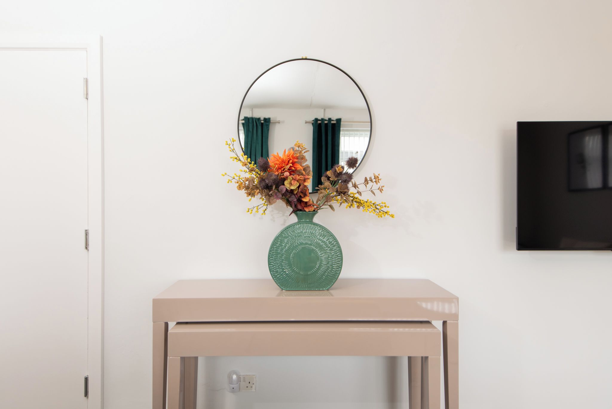 A room with a mirror on the wall, a vase with flowers on a table, and a door.