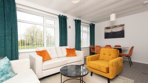 Un salon moderne avec un canapé blanc, un fauteuil jaune et une table basse ronde. On y trouve des rideaux verts, un coin repas avec des chaises orange et un tableau accroché au mur.