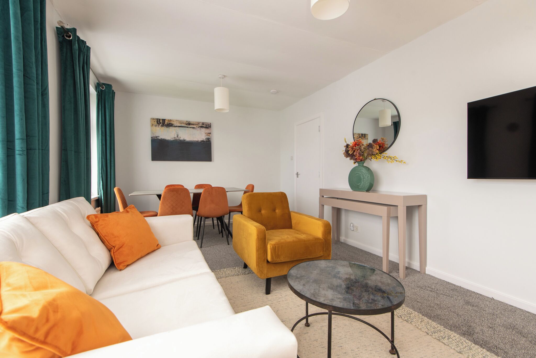 A modern living room with a white sofa, orange cushions, a yellow armchair, a round coffee table, and a dining area with orange chairs.