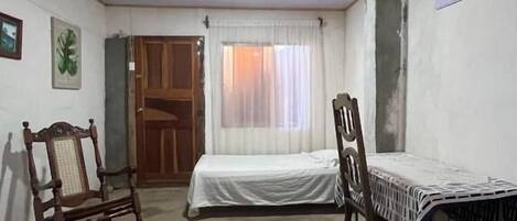 A room with a bed, two rocking chairs, a table with a patterned tablecloth, a window with curtains, and a door.