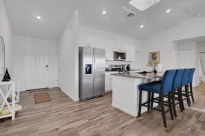 A modern kitchen with white cabinetry, a stainless steel refrigerator, a microwave, and a bar area with blue barstools.