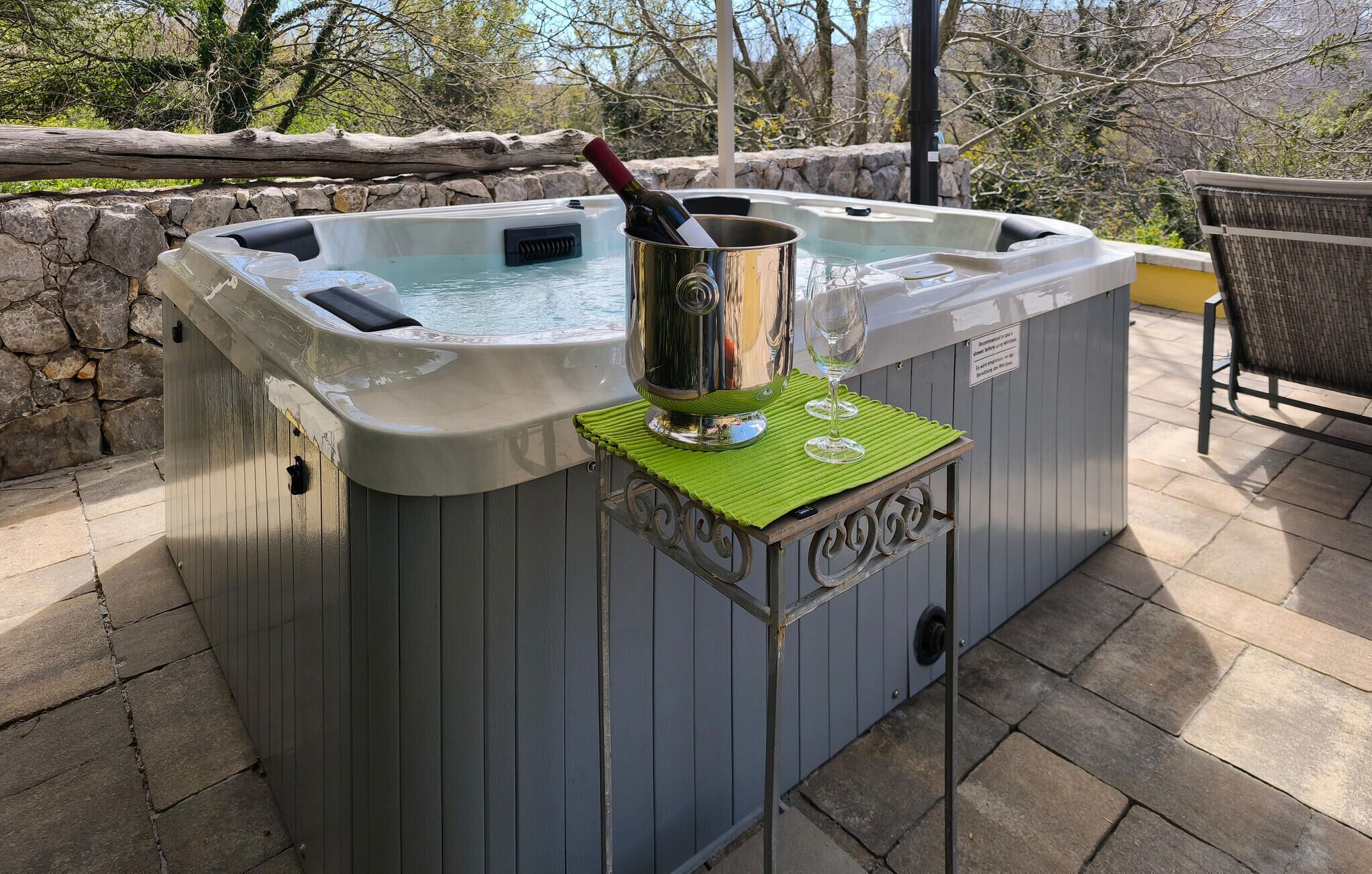 Un jacuzzi con una botella de vino y copas sobre una mesa, rodeado de paredes de piedra y mobiliario de exterior.