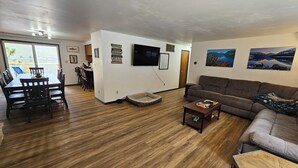 Een woonkamer met houten vloer, een grijze bank, een salontafel en een aan de muur bevestigde televisie. Er is een eethoek met een tafel en stoelen, en een klein hondenbed op de vloer.