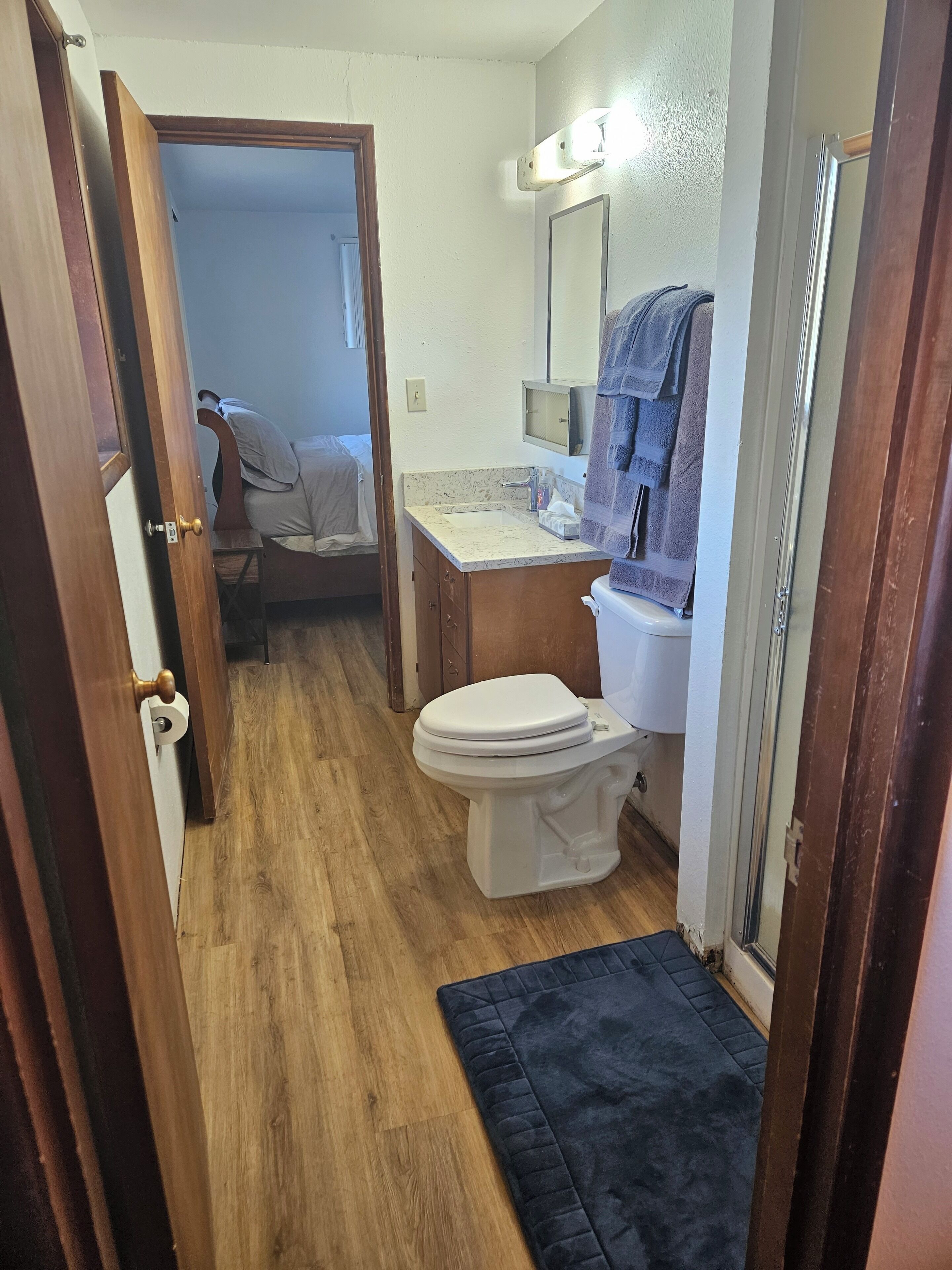A compact bathroom with a toilet, sink, and mirror. A door leads to a bedroom with a bed and a chair.