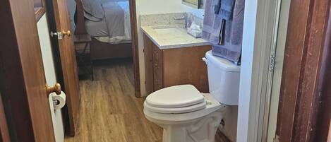 A compact bathroom with a toilet, sink, and mirror. A door leads to a bedroom with a bed and a chair.
