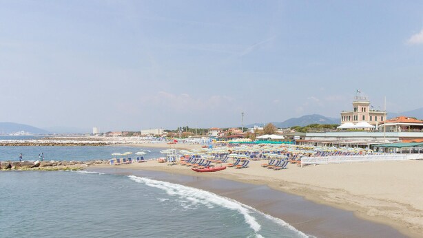 Una spiaggia con file di lettini e ombrelloni, un molo e un edificio con una cupola e una bandiera.