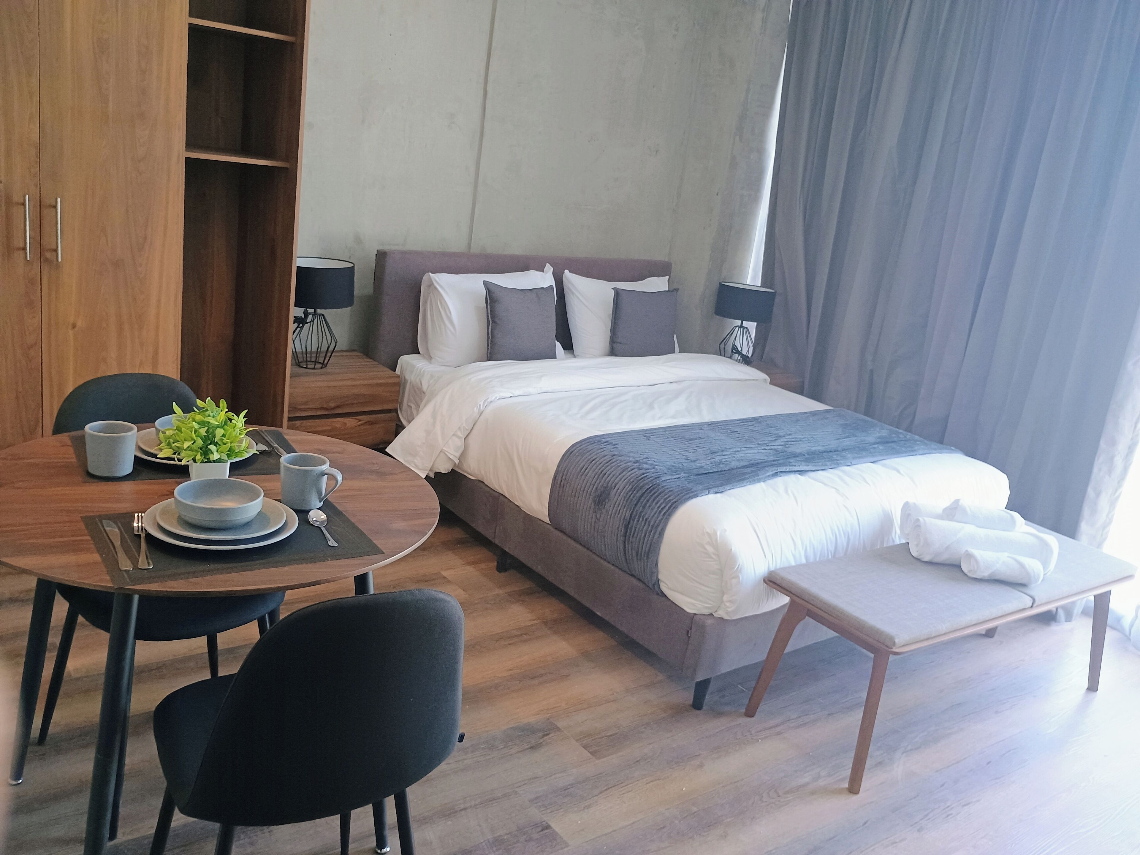 A hotel room with a bed, bedside tables, a dining table with chairs, a small plant, and a window with curtains.