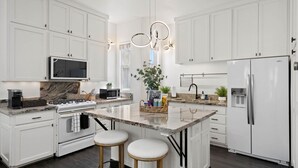 Cozinha moderna com armários brancos, uma ilha central com banquetas, forno embutido, micro-ondas, geladeira e luminária pendente.