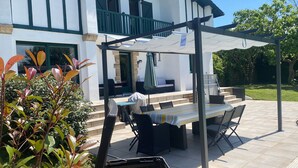 Un espace extérieur aménagé en terrasse, avec une table et des chaises, un coin salon abrité et une vue sur une maison moderne dotée d’un balcon.