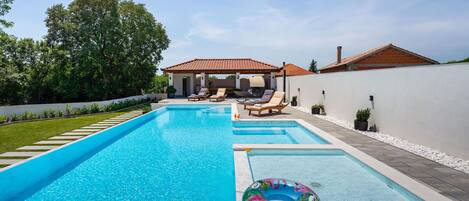 A rectangular swimming pool with clear blue water, surrounded by a white wall and a garden. There are lounge chairs and a covered seating area in the background.