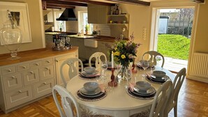 A dining area with a round table set for six, surrounded by white chairs. There is a kitchen in the background with a window and a view of a garden. - Beautiful 1700 century house in the heart of Viken. Pets Allowed! (Skåne län)