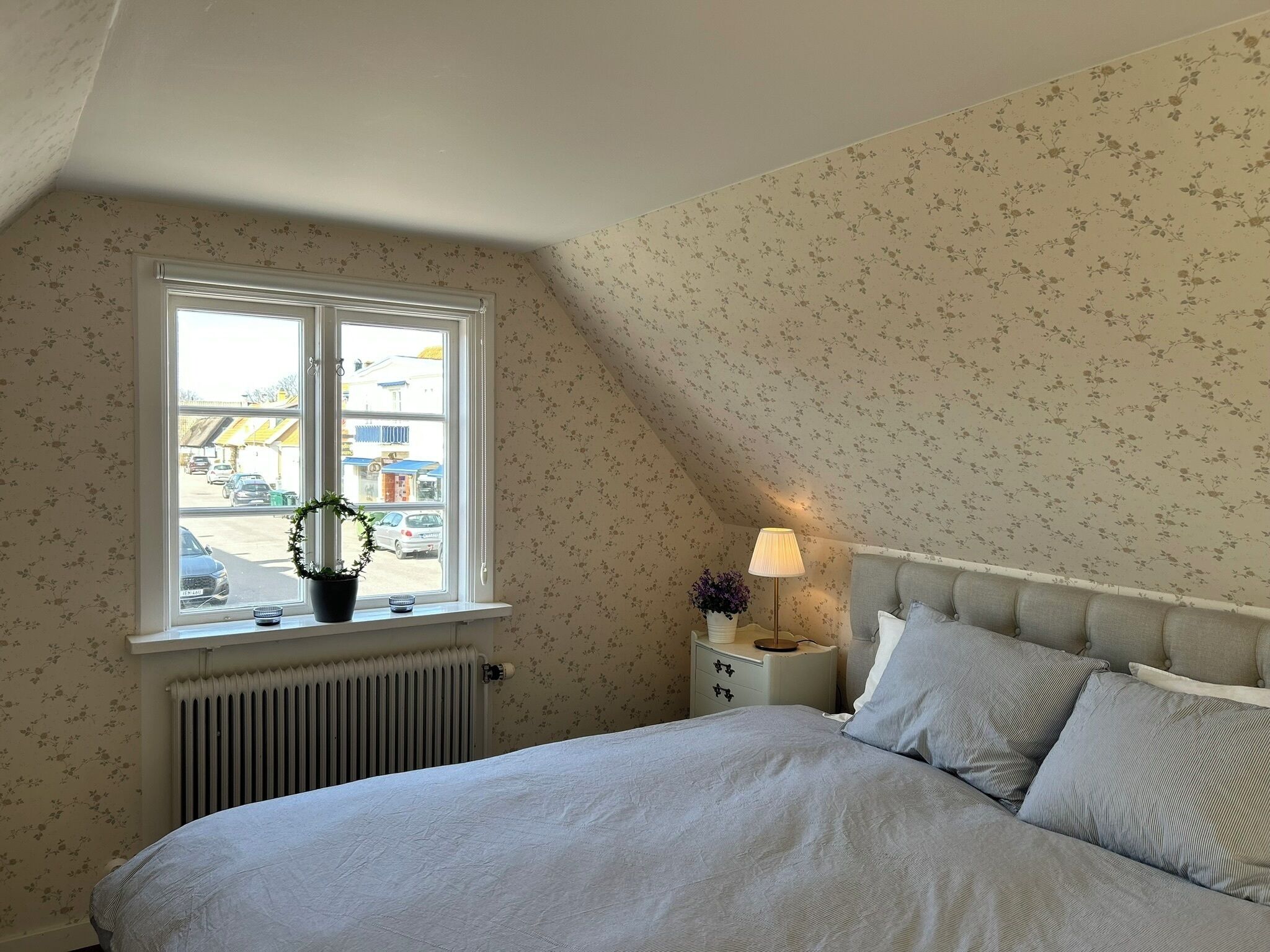 Een slaapkamer met een bed, nachtkastje, lamp, raam met uitzicht en bloemenbehang.