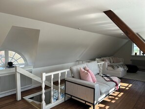 Un'ampia camera da letto in mansarda con soffitto spiovente, una scala bianca integrata, un divano grigio con cuscini rosa e una poltrona bianca.