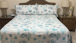 A bed with a blue and white ocean-themed bedspread, two bedside tables with lamps, and a wicker headboard. - PRICE REDUCTION!  Polynesian Village pool side! Walking distance to Pier Park (Panama City Beach)