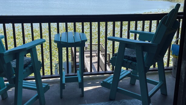 A balcony with two turquoise Adirondack chairs and a small table, overlooking a lake with lily pads and a distant shoreline with buildings. - PRICE REDUCTION!  Polynesian Village pool side! Walking distance to Pier Park (Panama City Beach)