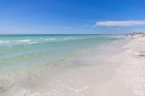 Une plage à l’eau turquoise et limpide, avec un rivage sablonneux et des bâtiments visibles au loin.
