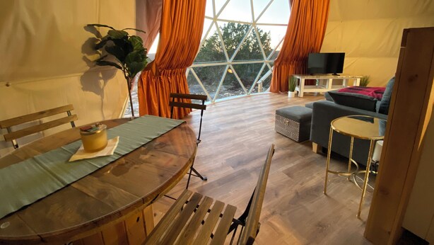 A tented accommodation with a wooden dining table, benches, a sofa, and a television. - Ventana Dome @ Rolling S Ranch (Bertram)