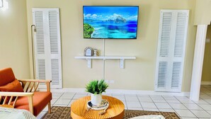 Een woonkamer met een aan de muur bevestigde televisie, een ronde houten salontafel met een plant, een bank en een fauteuil.