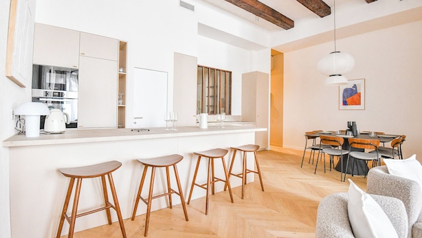 A modern kitchen with a central island, bar stools, a dining area with wooden chairs, and a large window. - Le Marcel - Appart standing 3 ch avec clim (Marseille)