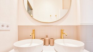 Une salle de bain avec deux lavabos blancs, des robinets dorés, un miroir rond et des articles de toilette de couleur beige.