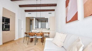 Un salon moderne avec un canapé blanc, un parquet en bois disposé en chevrons, un coin repas avec une table et des chaises en bois, et une cuisine équipée d'appareils électroménagers intégrés.