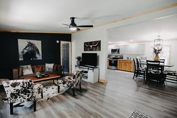 Una sala de estar moderna con un sofá, dos sillones, una mesa de centro y un televisor. Se observa un comedor con una mesa y sillas. La cocina es visible al fondo, con gabinetes y electrodomésticos blancos.