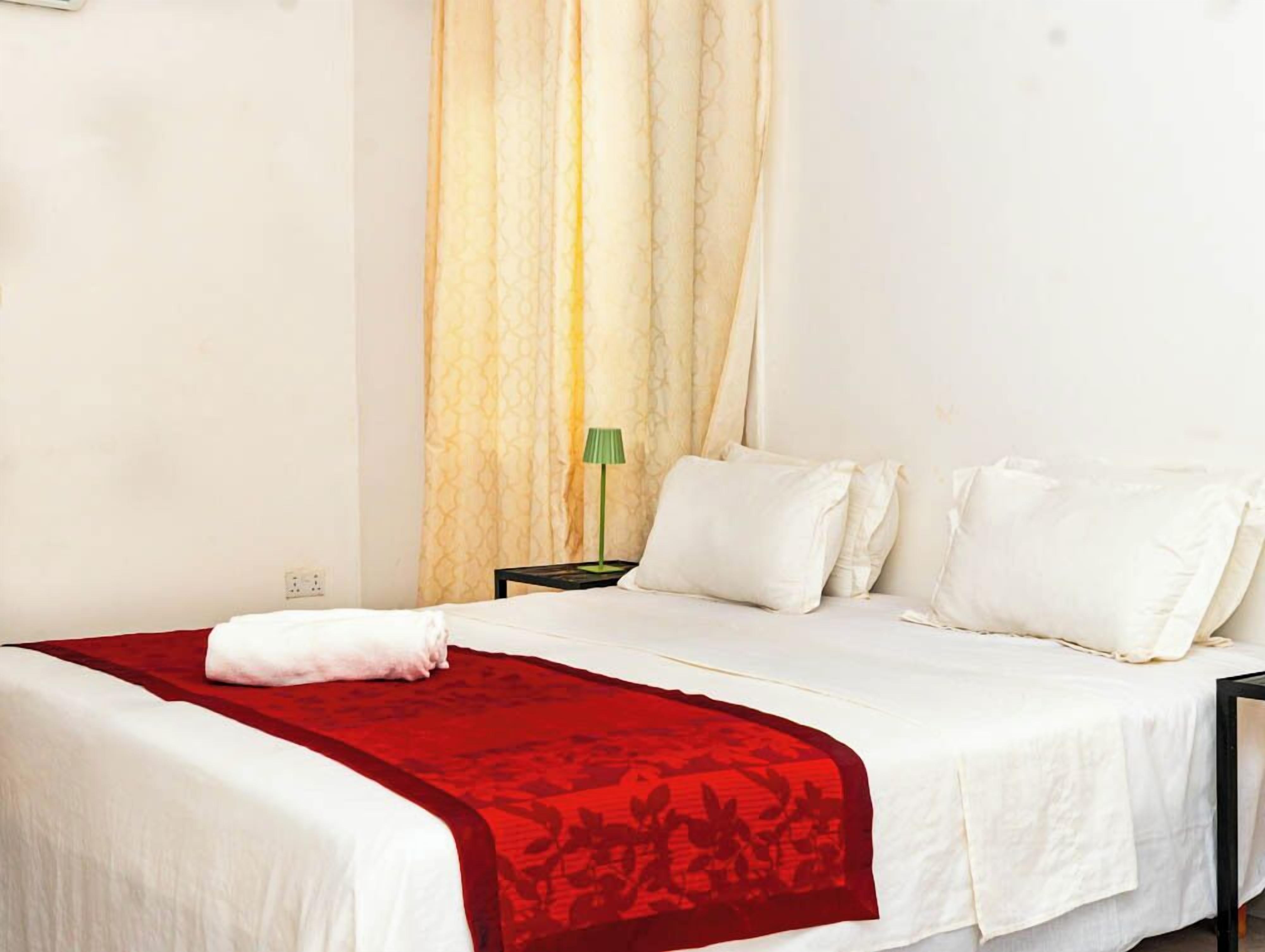Habitación de hotel con cama, ropa blanca, un plaid rojo, una lámpara verde y una cortina.