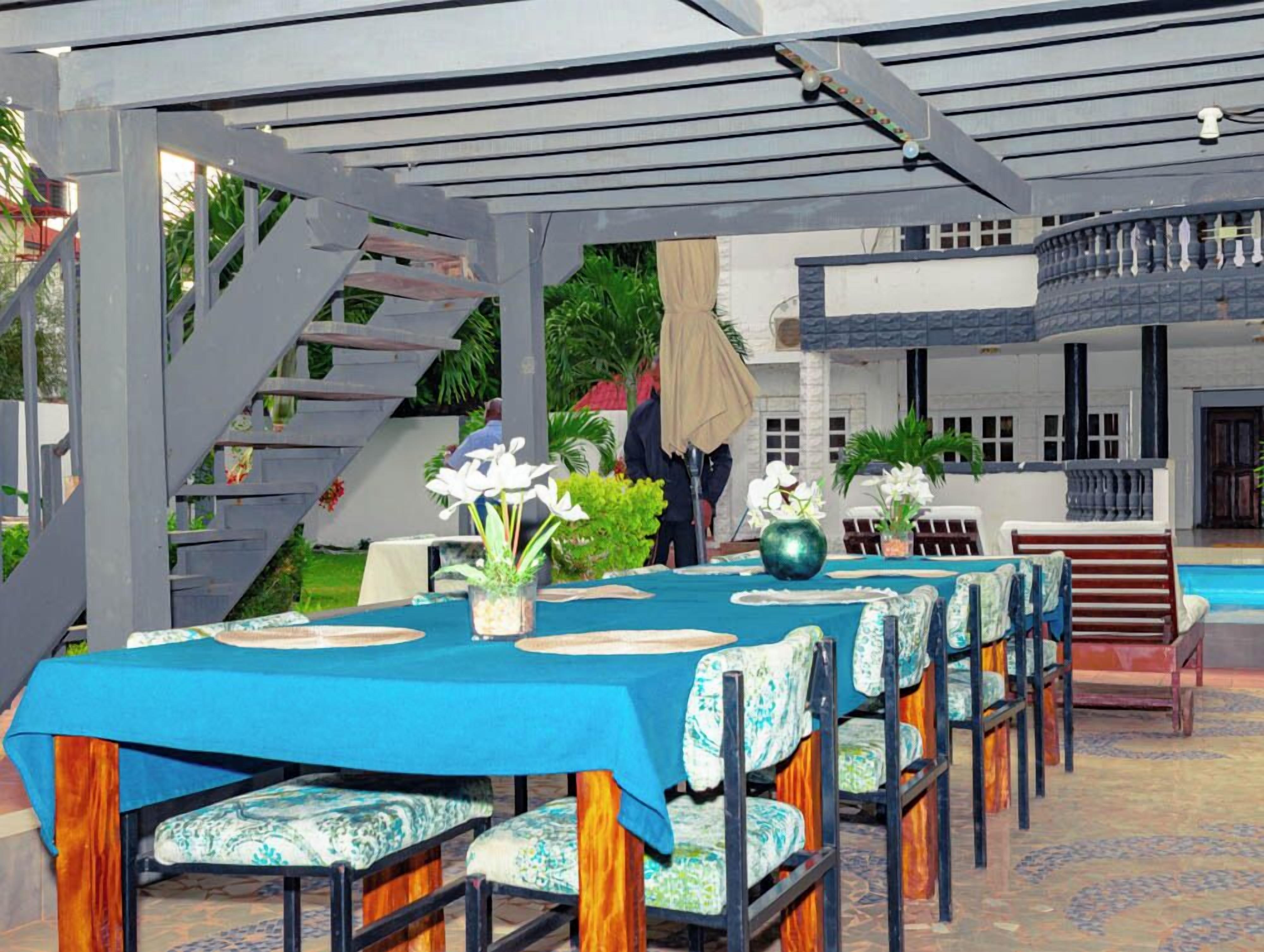 Un espace de restauration en plein air avec une nappe bleue, des chaises en bois et un escalier menant à un balcon.