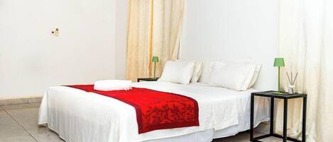 A hotel room with a bed, white curtains, a red bedspread, a green lamp, and a black nightstand.