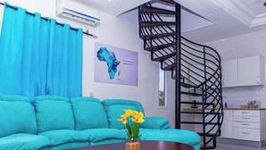 Ein Wohnzimmer mit einem türkisfarbenen Sofa, einem runden Couchtisch mit einer Vase voller gelber Blumen, einem blauen Vorhang, einer Karte Afrikas an der Wand und einer schwarzen Wendeltreppe.