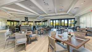 Un comedor amplio con varias mesas y sillas, ventanas grandes con vistas a zonas verdes y una barra central.