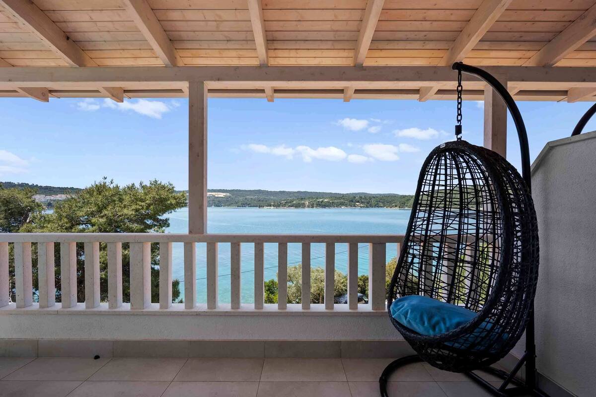 A wooden balcony with a hanging chair, a white railing, and a view of a body of water and distant landscape.