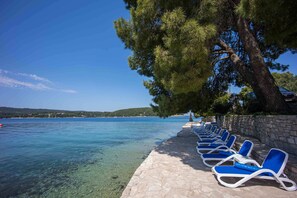 A row of blue lounge chairs by a stone-paved waterfront with clear water and a tree providing shade. - Boutique Guesthouse Sveti Petar (Solta)