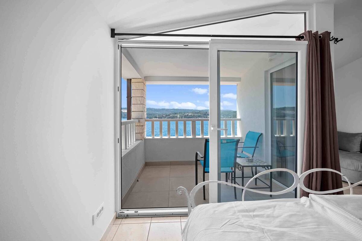 A bedroom with a bed, a glass door leading to a balcony with a view of the sea and a chair.