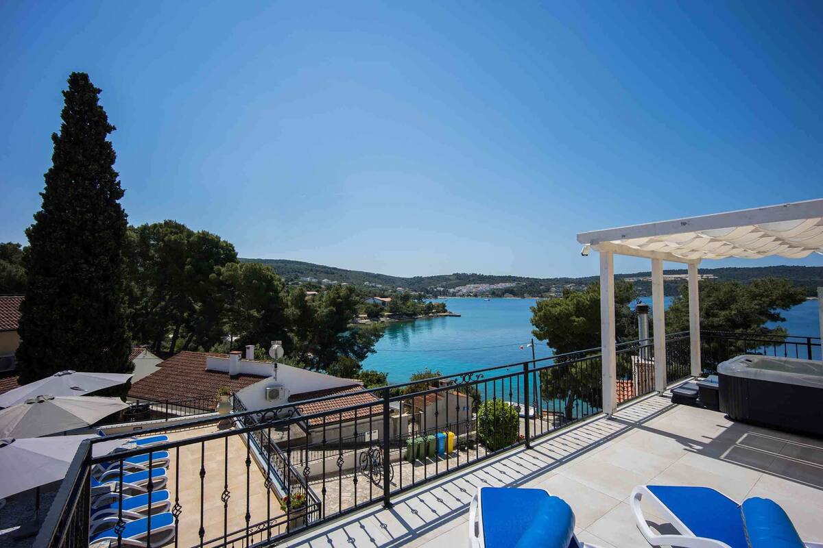 A balcony with blue lounge chairs, a pergola, and a view of a lake and hills.