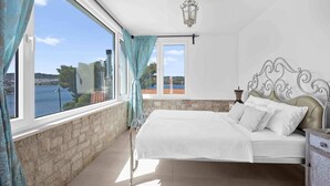 A bedroom with a bed, a nightstand, a lamp, and a view of a body of water through large windows. - Boutique Guesthouse Sveti Petar (Solta)