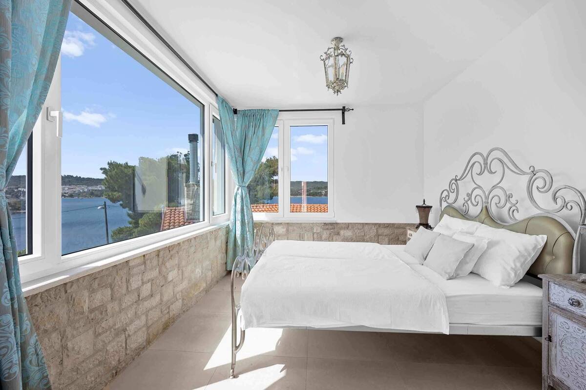 A bedroom with a bed, a nightstand, a lamp, and a view of a body of water through large windows.