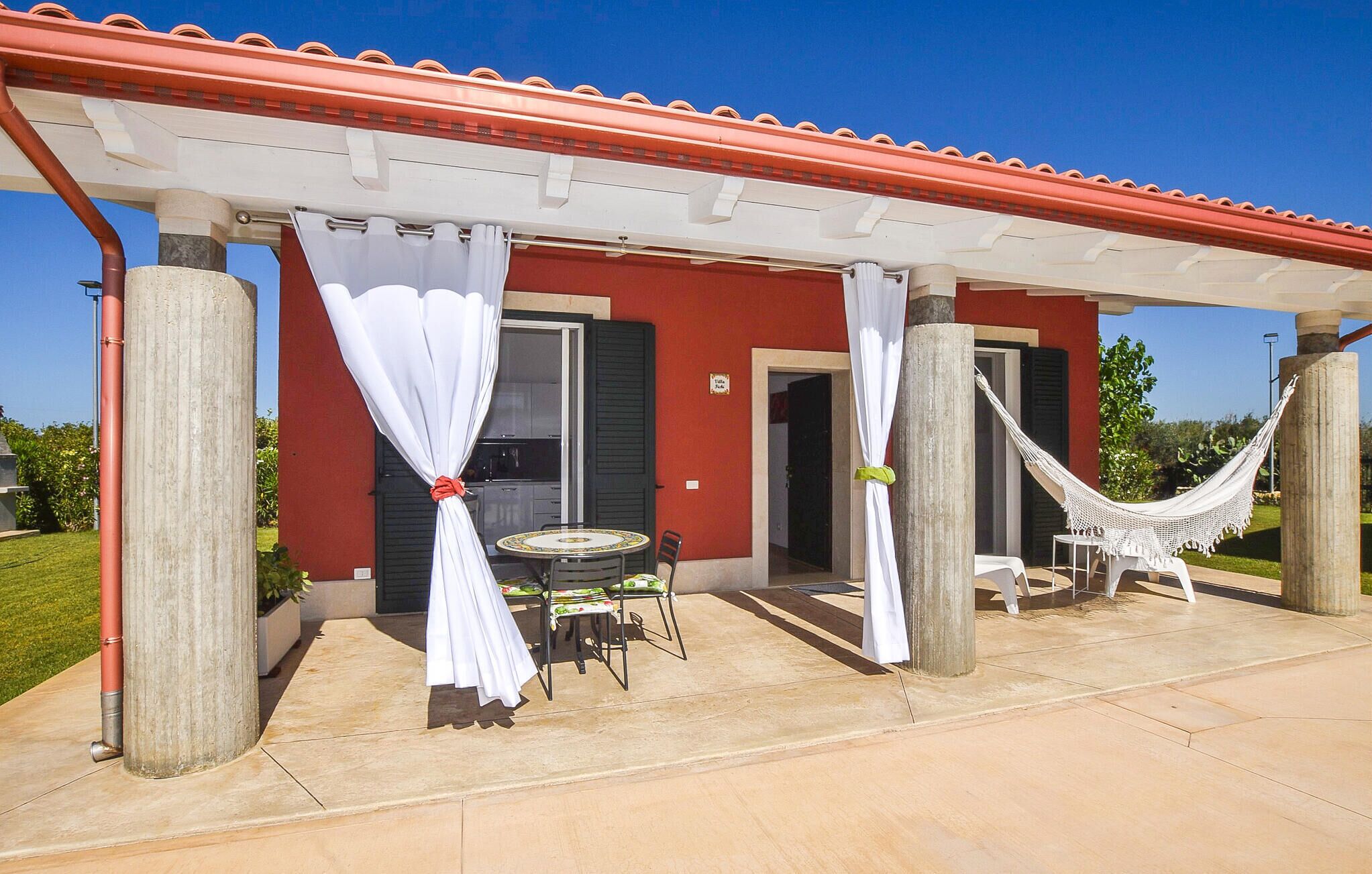 A covered outdoor area with a table and chairs, a hammock, and a red building with a tiled roof.