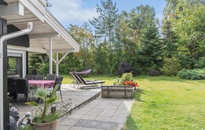 Un patio techado con mobiliario exterior, un jardín con diversas plantas y flores, y vistas de los árboles circundantes.