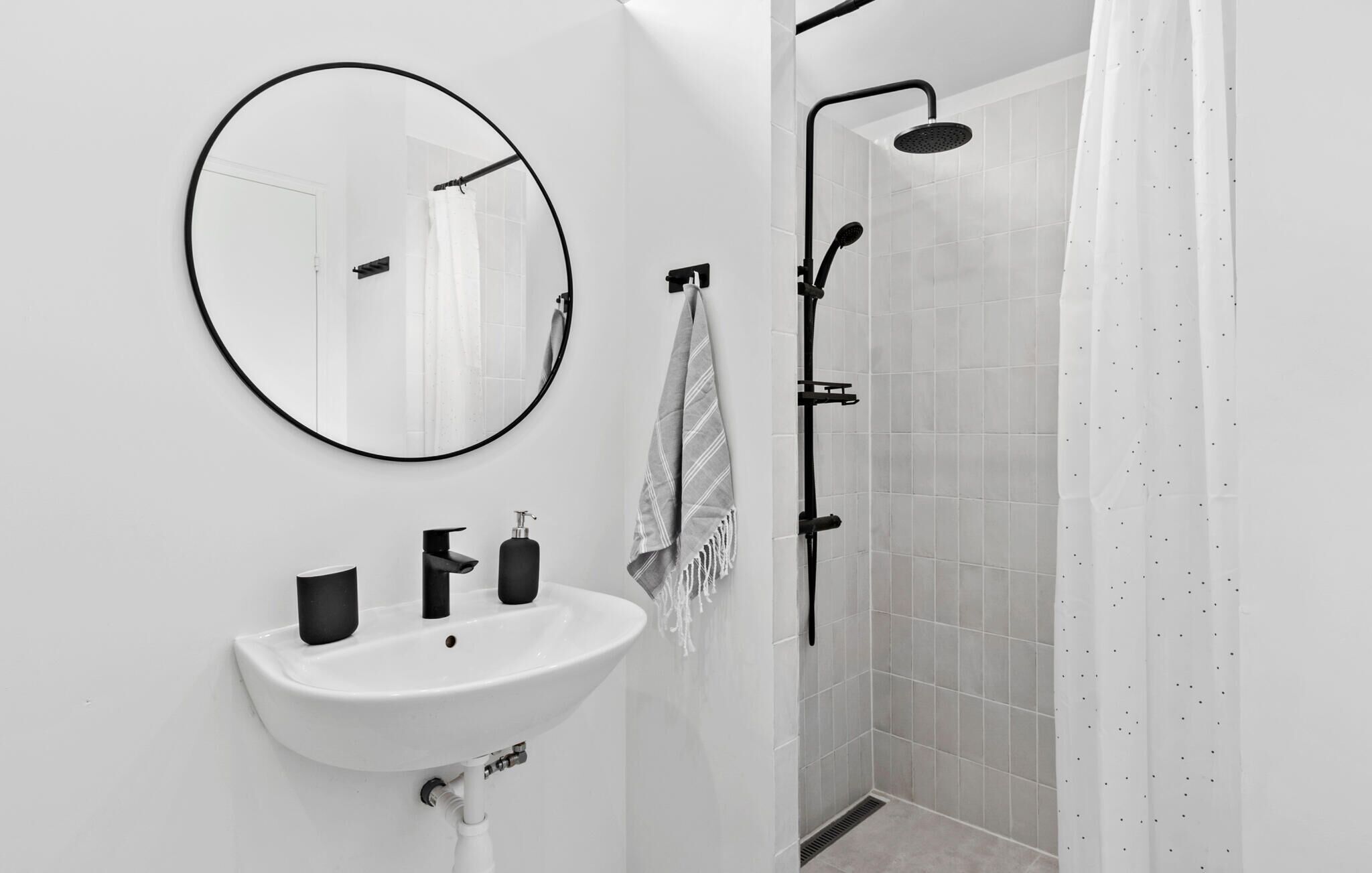 A modern bathroom with a round mirror, a white sink, black faucets, a towel, and a shower with a black hose.