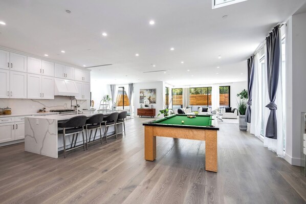 Una cocina moderna con armarios blancos, una zona de comedor con sillas, una mesa de billar y una sala de estar con sofá y una mesa de centro.