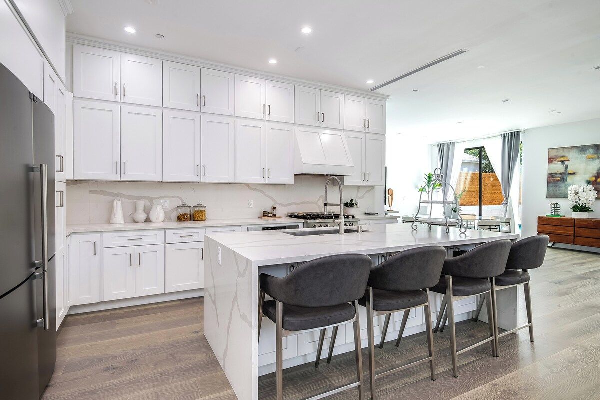 A modern kitchen with white cabinetry, a central island with bar stools, and a large window providing a view of the outdoors.