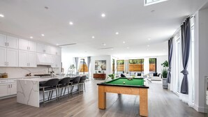 Une cuisine moderne avec des meubles blancs, un îlot central doté de tabourets de bar, une table de billard et un coin repas avec un canapé et des fauteuils.