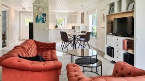 A modern living room with a sofa, a coffee table, and a TV. There is a dining area with a table and chairs. The room has a kitchen in the background.