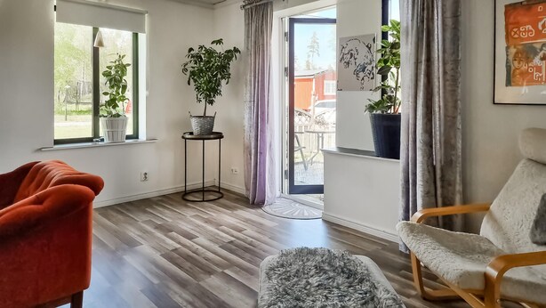 A modern living room with a wooden floor, a red chair, a grey ottoman, and a wooden chair. There are two windows with curtains, a potted plant on a small table, and a door leading to an outdoor area.