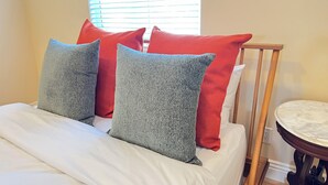 Ein ordentlich bezogenes Bett mit weißer Bettwäsche und zwei grauen Kissen, einem roten Kissen sowie ein kleiner runder Tisch mit Marmorplatte daneben.