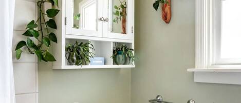 Une salle de bain comprenant des toilettes, un meuble blanc avec des miroirs, une étagère avec des plantes et un porte-rouleau de papier toilette.